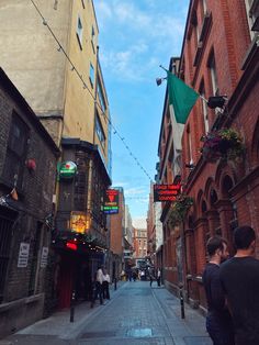 two men are walking down an alleyway in the middle of town on a sunny day