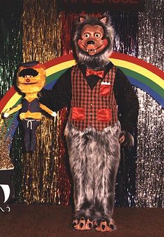 a man in a bear costume standing next to a teddy bear with a rainbow behind him