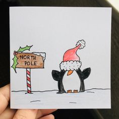 a hand holding up a christmas card with a penguin wearing a santa hat next to a sign that says north pole