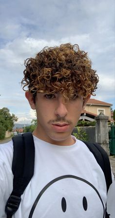 a young man with curly hair and a backpack looks at the camera while standing in front of a house