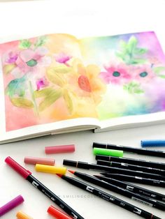 an open book with watercolor pencils next to it and some flowers on the page