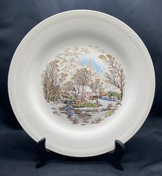 a white plate with a winter scene painted on the front and sides, sitting on a black stand