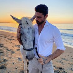 a man standing next to a white horse on top of a sandy beach near the ocean