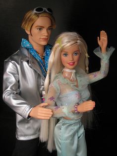 two barbie dolls are posed next to each other, one is wearing a silver jacket