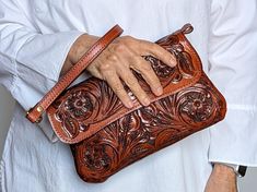 a woman's hand holding a brown leather purse with floral designs on it, while wearing a white shirt