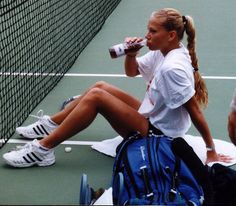 a woman sitting on the tennis court drinking from a bottle while holding a backpack and water bottle