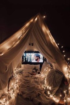 a bed covered in white sheets with fairy lights on the top and bottom, next to a flat screen tv