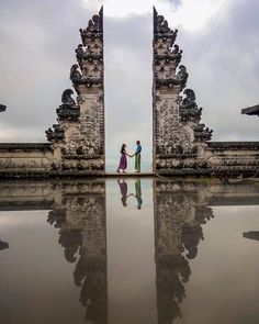 two people are standing in front of an ornate gated entrance to a temple with water reflecting on the ground