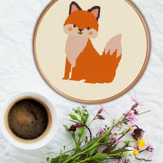 a cross - stitch fox sitting next to a cup of coffee and some wildflowers