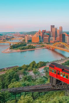 a red train traveling over a bridge next to a large city on top of a lush green hillside