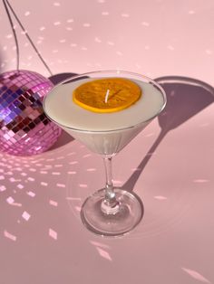 an orange slice sitting in a martini glass with a disco ball behind it on a pink surface