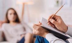 a woman sitting on a couch while holding a pen and paper in her hand, with another person behind her