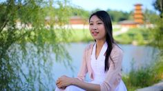 a woman sitting on the ground next to a body of water with willow trees in the background