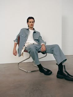 a man sitting in a chair with his legs crossed and wearing black boots, looking at the camera