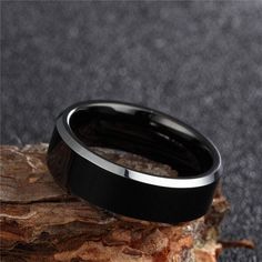 a black and silver wedding ring sitting on top of a piece of wood with a rock in the background