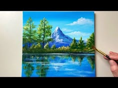 a person is holding a paintbrush in front of a painting of a mountain lake