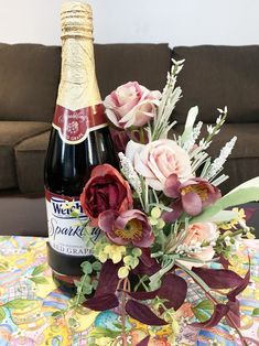 a bottle of wine and some flowers on a table with a couch in the background