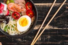 a bowl with noodles, meat and vegetables in it next to chopsticks on a wooden table