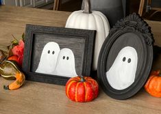 two framed pictures with ghost faces are sitting on a table next to pumpkins and gourds