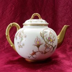 a white tea pot with gold trimming and flowers painted on the side, sitting on a pink background