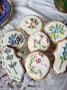 several decorated cookies are on a glass platter with lace doily and blue flowers