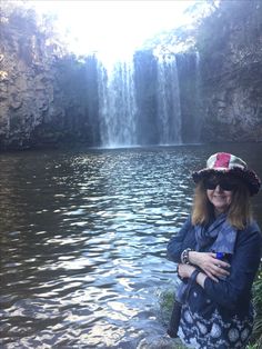 a woman wearing a hat standing in front of a waterfall with her arms around her chest