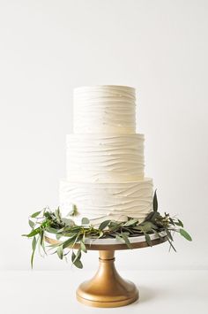 a white wedding cake with greenery on the top and bottom is sitting on a gold pedestal