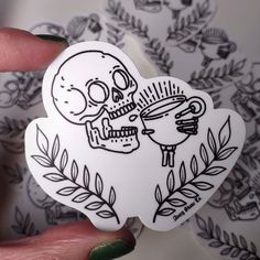 a hand holding a sticker with a drawing of a skull and a cup on it