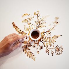 a person is drawing on a wall with gold foil flowers and leaves, while holding a coffee mug