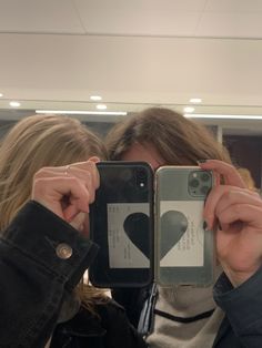 two women taking pictures with their cell phones in the mirror, one holding up her phone