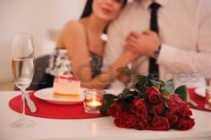 a man and woman sitting at a table with wine glasses, roses and cake on it