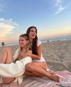 two women are sitting on the beach and one is wrapped in a towel while the other smiles