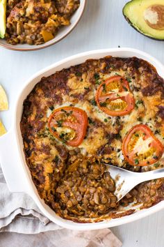 a casserole dish filled with beans, tomatoes and cheese next to avocado