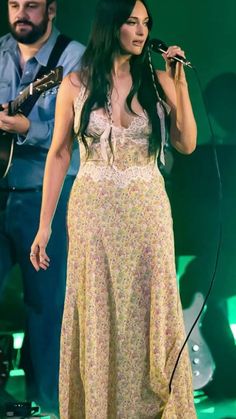 a woman in a long dress holding a microphone and standing next to a man with a guitar