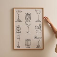 a woman is holding up a framed poster with cocktails on the wall behind her