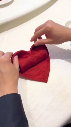 a person is making a heart out of red material on a white tablecloth with wine glasses and plates in the background