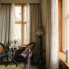 two chairs and a table in front of a window with drapes on the windowsill