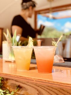 two cocktails sitting on top of a wooden table in front of a woman at a bar