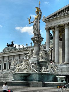 a statue in front of a building with columns and statues on the outside, along with people walking around