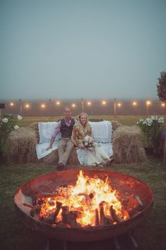 two people sitting next to each other in front of a fire pit with hay bales