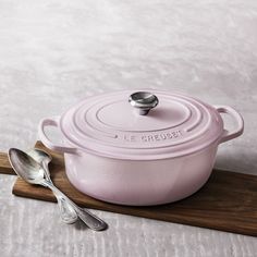 a pink casserole on a wooden tray with two spoons and a serving utensil