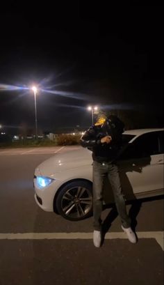 a man standing next to a white car in a parking lot at night with the lights on