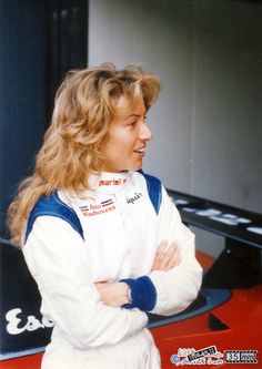 a woman standing in front of a red car with her arms crossed and looking off to the side