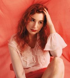 a woman sitting on top of a red sheet with her hands behind her head and looking at the camera