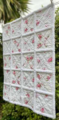 a white quilt hanging from a clothes line with pink flowers on it and green bushes in the background