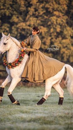 a woman riding on the back of a white horse in a field with trees behind her