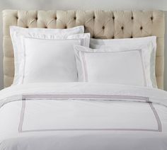 a bed with white sheets and pillows on top of it, in front of a beige headboard