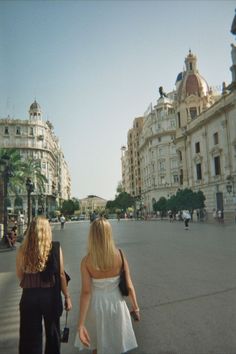 two women are walking down the street in front of some buildings and one is carrying a suitcase