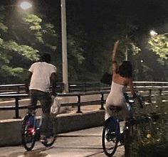 two people riding bikes down a sidewalk at night with their arms up in the air