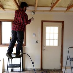 a man standing on a step ladder painting the ceiling in an unfinished room with two windows